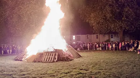 Shaun Hennessy A group of people gather on the edges of a green near houses to watch a bonfire made using wood and old pallets