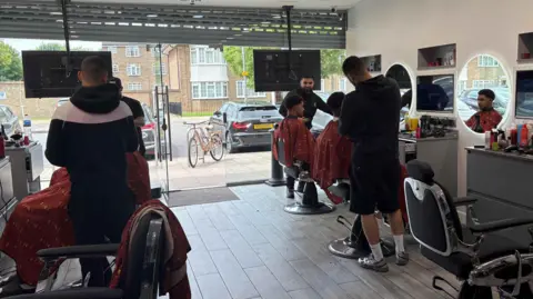 Four men are sitting in chairs in a barber salon as hair stylists work on their cuts. The customers face circular mirrors and grey cabinets as they get their hair done