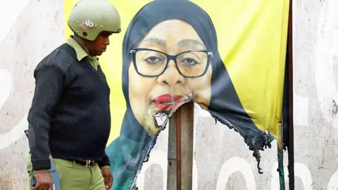 Reuters A Tanzanian riot police officer walks past a vandalised campaign poster of President Samia Suluhu Hassan showing her dressed in a black headscraf against a yellow background - 30 October 2025.