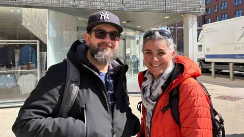 A man and a woman are stood together smiling towards the camera. The man has his hands in the pockets of his black jumper with a backpack on and he has a beard and is wearing a black cap and sunglasses. The women is wearing a red jacket and a white scarf with sunglasses on her head.