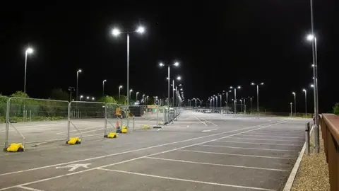 Mike Barrett A large car park with multiple street lights, shown at night with the lights on and a barrier down the middle