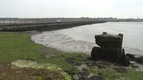 Neil Theasby/Geograph.org A single white car can be seen driving along a fenced causeway above tidal mudflats.