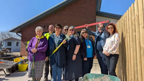 Members of the four different charities, they smile and group together, one has a hammer, someone else holds a plank, there is a red building in the background.