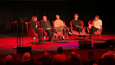 Five people sit on red chairs on a stage - making up a panel - with an audience looking on.