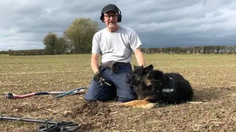 Chris Revill A man in a grey t-shirt, navy trousers, black cap and headphones is crouching in a field. He has a tool in his hand and a black dog laid beside him. There is a spade and a metal detector also laid on the ground. There are hedgerows and trees in the distance. The sky is cloudy.