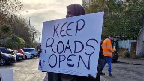 A woman protestor in Barton Hill holds a sign saying 'Keep Roads Open'. She is standing in a road with a man in an orange high vis jacket in the background. 