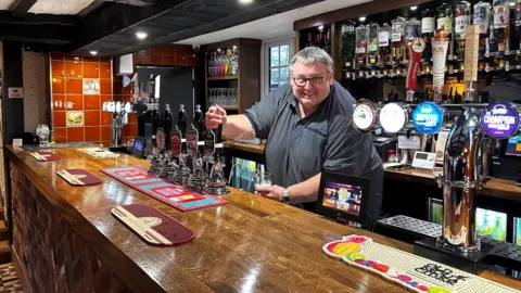 Darren is behind the bar of the his pub pulling a pint and smiling at the camera. He has a grey T-shirt on and glasses with short, brown hair. Drinks can be seen on shelves behind him.