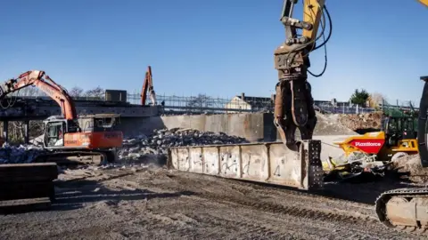 North Somerset Council A large machinery arm picks up a metal piece of a demolished bridge on a demolition site. Other construction vehicles can be seen moving rubble around the site. A temporary footbridge, replacing a gap where the bridge once stood, can be seen in the distance, and it is a sunny winter day with clear skies.