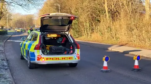 Owen Ward/BBC A police vehicle and cones blocking off a road. There is frost on the grass on a verge on on the left, and trees bathed in sunshine on the right. 