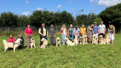 A row of 13 people standing and crouching in a line, with their golden retrievers at their feet.