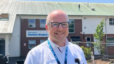 A man with thinning hair smiles at the camera as he stands outside a surgery on a sunny day. He is wearing glasses and an NHS lanyard.