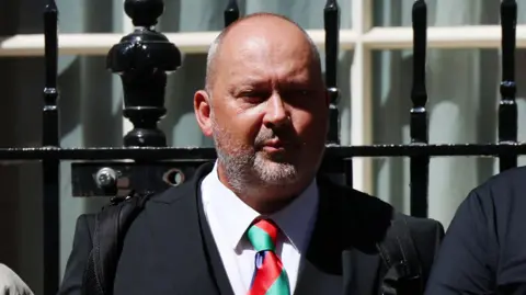 Olly Stephens' father Stuart stands outside 10 Downing Street. He is wearing a dark suit and a red and green tie.