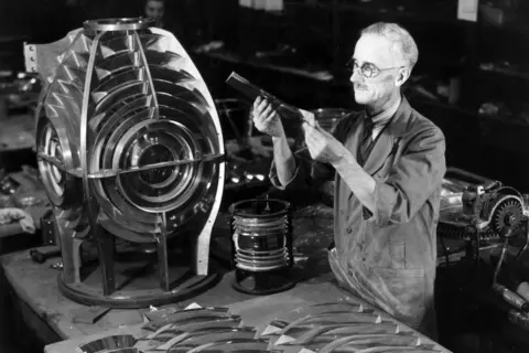Getty Images A black and white image of a glass factory worker. He is wearing round rimmed glasses and inspcting a piece of glass. In front of him is the metal casing for a lighthouse. 