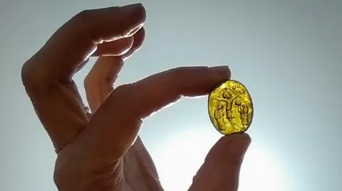 Oxford Cotswold Archaeology Medieval Venetian glass pilgrims token held up to the light between a thumb and forefinger. It is golden yellow in colour and oval shaped. It shows a crucified Christ with two figures with bowed heads on either side of him.