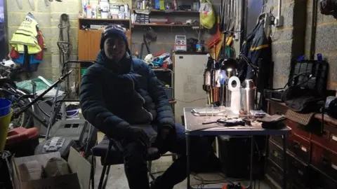 Adam Root Adam Root working in a dark garage surrounded by clutter. He is wearing a hat, coat and gloves and is sitting at a desk.
