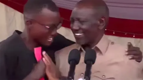 A blurry screengrab from the incident showing a man in glasses, holding a book, putting his arm around William Ruto. Ruto is smiling, but appears to be pushing him away.