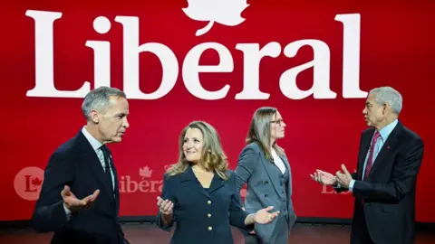 Mark Carney, Christia Freeland, Karina Gould and Frank Baylis stand speaking to one another in front of a "Liberal" sign while attending a leadership debate in Montreal in February.