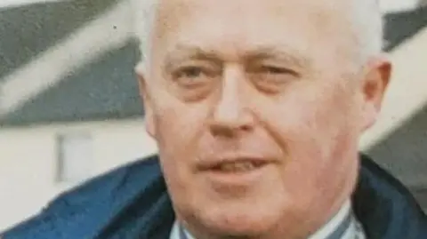 Ivan Barr has short grey hair and is wearing a chequered grey shirt. He is outside near a black fence and a number of trees. 