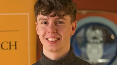 Dr Jack Devin is smiling at the camera. He has dark hair with a long curly fringe and short sides. He is wearing a polo neck jumper.