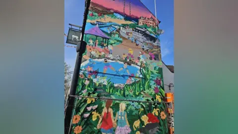 Wychavon District Council The side of a building completely covered in a mural. It depicts a street scene in bright colours, with houses, a street and a canal. There is also a bandstand with people playing instruments, two people sitting in a pool and vibrant greenery with animals including otters and bright flowers. Two girls are holding hands at the bottom, looking over the rest of the mural.