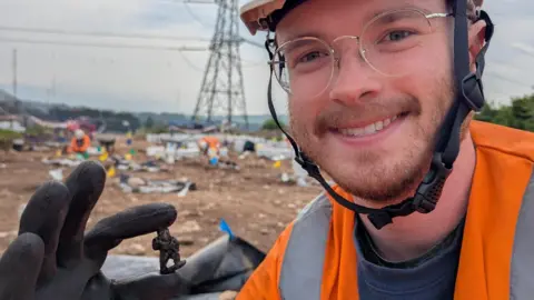 Man with white hard hat, glasses and beard, smiling and wearing an orange hi-vis top and black gloves holding a dark figurine. He is on a burial site with a pylon in the background.