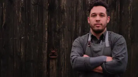 Abbie Morris Corrin Harrison in grey apron and dark grey shirt and arms crossed. He is standing in front of a wooden door and has dair hair and short trimmed dark facial hair