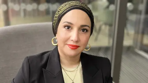 Headshot of Reem sitting down, wearing a headscarf and black jacket.