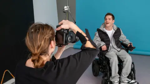 BBC A man, Elliot Caswell, is sitting in an electric wheelchair in front of a blue backdrop in a photography studio. He is wearing a grey tracksuit and a black gilet and is smiling at the camera. A woman in a black top is taking his photo with a large camera.