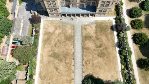 A parched lawn with historic patterns against a stately home