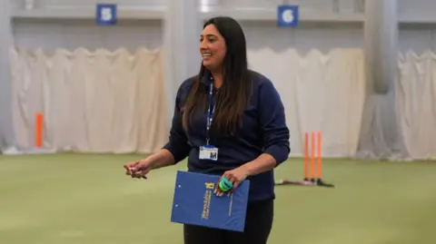 Warwickshire Cricket Foundation Simran is wearing a navy blue long-sleeved sports polo top and matching trousers. She has a navy blue lanyard around her neck and is holding a clipboard that has the yellow Warwickshire Cricket Foundation logo printed on the back of it. In the other hand she is holding a pen and she has a french manicure. She is smiling with her teeth and looking off to the side. Simran has long, dark brown hair. The background is out of focus, but there are white sheets and netting, as well as orange plastic cricket stumps on the green astro floor.