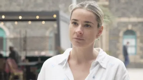 Zara pictured with the Pavilion restaurant visible in the background, though it is slightly blurred. Zara has her blonde hair in a ponytail and is wearing a white collared shirt. 