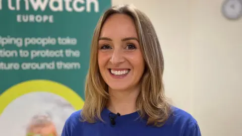 Kate Bradbrook/BBC Dr Sasha Woods is in the middle smiling at the camera. She has shoulder length light brown hair and brown eyes, and is wearing a blue T-shirt. She is stood next to a sign on the left. 