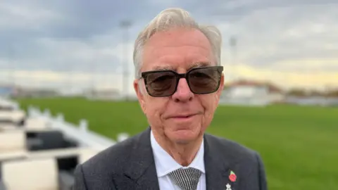 Philip Miller stands outside on a cloudy day in front of a large section of green grass. He has grey hair and wears sunglasses, a grey suit jacket, white shirt, black and white tie, and a small poppy pin on one side of his chest. 