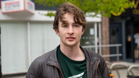 Alex looking at the camera, stood in Bangor town centre in front of an office building. He wears a brown leather jacket, green t-shirt and has short brown hair. 