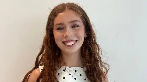 Grace Warren Grace heeft lang bruin haar en draagt ​​een top met stippen. Ze staat voor een witte achtergrond en glimlacht.