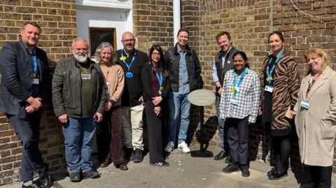 West Hertfordshire Teaching Hospitals NHS Trust A group of people gather around a grey plaque to commemorate former residents of the Watford Union Workforce. It's a sunny day and their standing in front of brick walls.