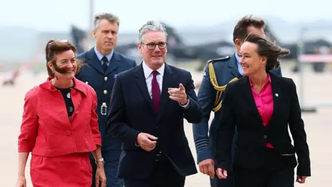 Getty Images UK Prime Minister Keir Starmer (C) arrives with Wales' First Minister Eluned Morgan (L) and Britain's Wales Secretary Jo Stevens (R) for a visit to RAF Valley. Morgan wears a pink jacket with a pink skirt. Sir Keir wears a dark suit with a red tie. Stevens wears a dark suit with a bright pink shirt.