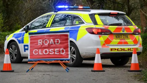 A stationary police car with its blue lights flashing. In front of it is a road closed sign and three traffic cones.