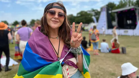 Taneesha stands outdoors at an event, draped in a rainbow flag and making a peace sign with her right hand. In the background, people are gathered on the grass near a stage and tents under a partly cloudy sky.