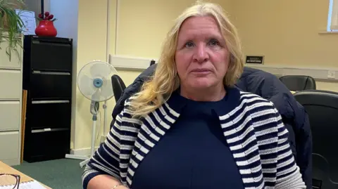 Christine Nicholls, wearing a blue and white top in front of a laptop looking straight at the camera.