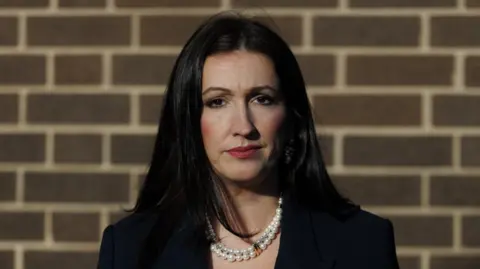 Emma Little-Pengelly, with long black hair, wearing a black jacket. She is standing in front of a brick wall. 