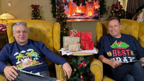 Two men wearing pirate themed Christmas jumpers sit in yellow arm chairs holding paper and pens. There is a table in the middle with Christmas presents on it and Christmas wreaths around them. A screen in the background shows a fireplace.