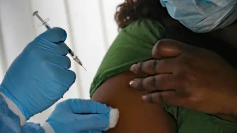 A person is shown receiving a flu vaccine in their upper right arm. A person can be seen holding a needle in a blue gloved hand and dabbing a spot on the patient's arm with a cotton bud. 