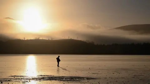 Georgina Maxwell A frozen loch with the sun splitting through the clouds. A figure in silhouette is making their way across the loch. 