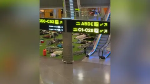 Ian Scott Passengers lying on the floor at Doha Airport. Arabic signage can be seen directing people to gates. Many of the passengers have blankets on them.