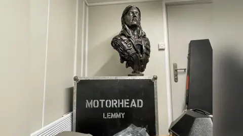 A bust of Lemmy with his ashes on a suitcase with the inscription "Motörhead Lemmy"