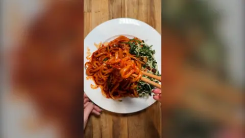A white plate with noodles in a tomato sauce being picked up by a pair of chopsticks. Two hands are seen either side.
