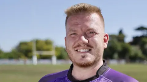 A man - Sam Biscoe - in a purple rugby top smiling at the camera. He is standing in front of a rugby pitch.