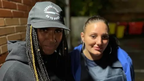 Danyelle King is on the left and has long braids, a black cap and is wearing a black hoodie. Jordan is on the right and her hair is slicked back into a ponytail and she is wearing a blue hoodie and a stab-proof vest.