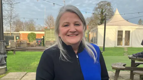 BBC/Amy Holmes Anne-Marie Vlader has light blonde hair and is wearing a dress with blue and black squares on it. She is sat down with her hands on the table in a pub garden